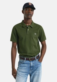 Jonge man in een donkergroen polo-shirt, lichtblauwe jeans, bruine riem en een marineblauwe honkbalpet met een "G" embleem, staande met één hand in zijn zak.