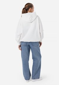 Felpa bianca con cappuccio e spalle scese, abbinata a jeans larghi azzurri. Il modello indossa sneakers bianche. Vista da dietro.
