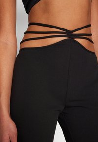 Mustad leggingid sujuva tekstuuriga on varustatud mitmete mustade rihmadega, mis ristuvad vööl, luues ainulaadse disainielemendi.