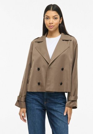 Kvinde med langt mørkt hår iført en kort brun dobbeltradet jakke, hvid top og blå jeans, der står mod en ensfarvet baggrund.