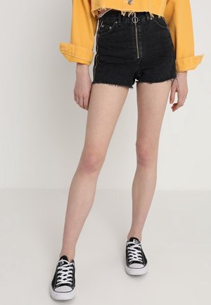 Person iført sorte denim shorts med lynlås foran, gul cropped jakke og sorte lave sneakers med hvide snørebånd.