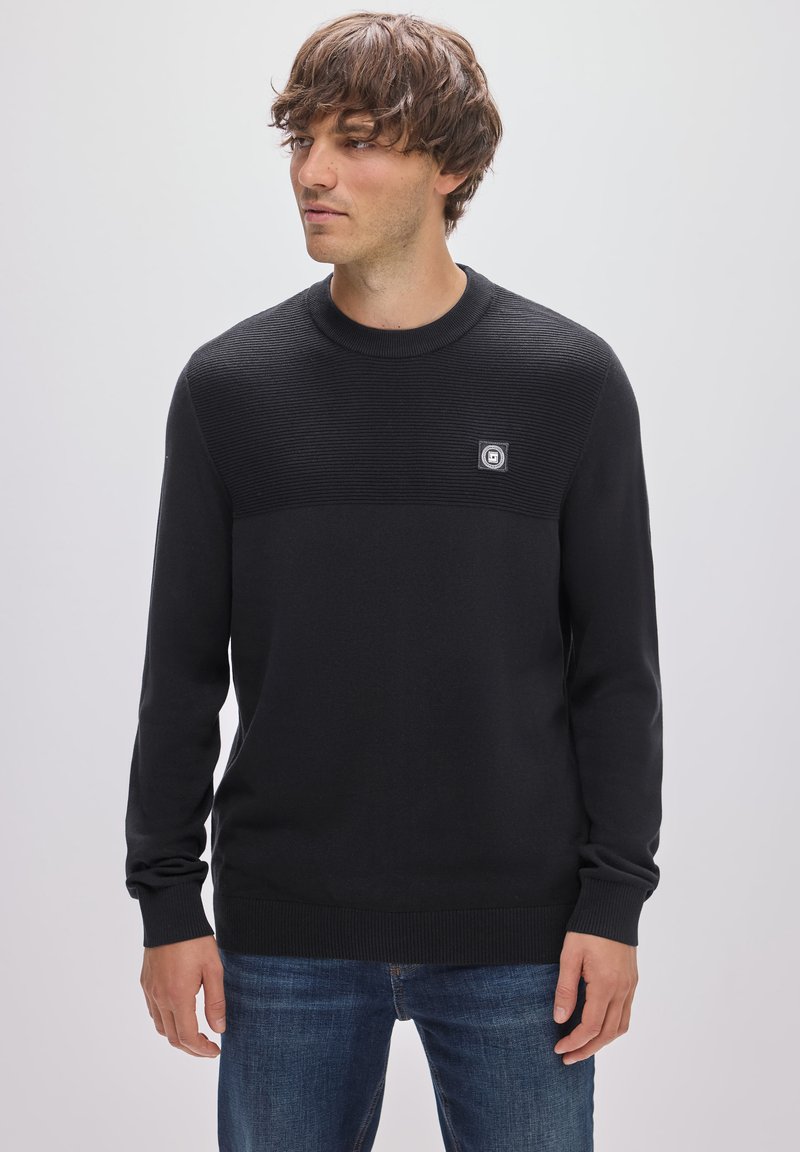 Schwarzer Pullover aus weichem Strickstoff mit einer gerippten Struktur im oberen Bereich und einem kleinen Logo-Patch auf der Brust.