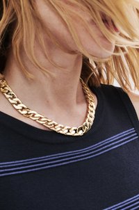 Goldene Kettenhalskette mit ineinandergreifenden Gliedern, getragen über einem schwarzen Oberteil mit drei horizontalen lila Streifen. Nahaufnahme.