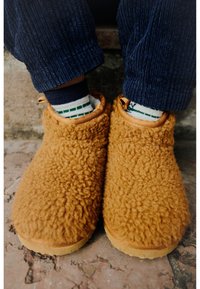 Καφέ, γούνινες παντόφλες με πλούσια υφή και στρογγυλό σχήμα. Διαθέτουν αντίθεση στην εξωτερική σόλα και συνδυάζονται με ριγέ κάλτσες.