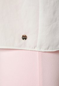 Tissu blanc avec une petite étiquette ronde en argent attachée près de l'ourlet sur un fond textile rose clair.