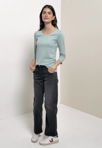 Femme debout, les mains dans les poches, portant un haut à manches longues bleu clair, un jean noir et des baskets blanches avec un détail rouge.