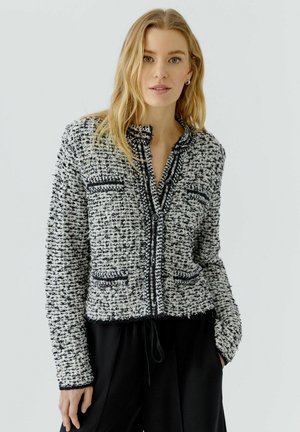 Vrouw met blond haar die een zwart-witte, gestructureerde jas met knopen en een zwarte broek draagt, staand tegen een effen lichte achtergrond.