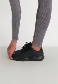 Scarpe da ginnastica nere con tomaia in rete, lacci e suola in gomma, abbinate a leggings grigi a coste attillati, mani che aggiustano la scarpa.