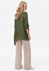 Oliwkowy, oversize sweter dzianinowy z podwiniętymi rękawami i teksturowanym wzorem, połączony z jasnobeżowymi spodniami o szerokich nogawkach i białym obuwiem.