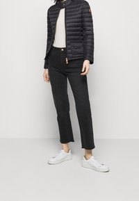 Une personne se tient debout, portant une doudoune noire, un haut beige et un jean noir coupé, associés à des baskets blanches, devant un fond neutre.