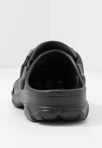 Sabot noir à enfiler avec semelle texturée et bride réglable, vu du talon sur un fond blanc uni.