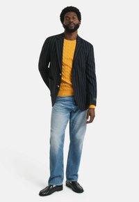 Blazer met marineblauwe streepjes, gouden knopen, gecombineerd met een gele kabelgebreide trui, lichtblauwe jeans en zwarte schoenen.