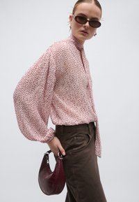 Ροζ πουκάμισο με σχέδια και φουσκωτά μανίκια, συνδυασμένο με καφέ παντελόνι. Αξεσουάρ: μικρή τσάντα με υφή κροκοδείλου σε βουργουνδί απόχρωση.