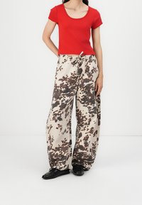 T-shirt rouge ajusté avec un col dégagé, associé à un pantalon large beige à motif tacheté marron et une taille élastique, porté avec des chaussures noires.