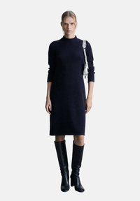 Abito in maglia a maniche lunghe di colore navy con collo alto, abbinato a stivali neri al ginocchio e una piccola borsa a tracolla in argento.