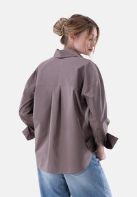 Camicia a maniche lunghe in morbido tessuto taupe, con un plissé sul retro, colletto classico e maniche con polsini, abbinata a jeans blu chiaro.