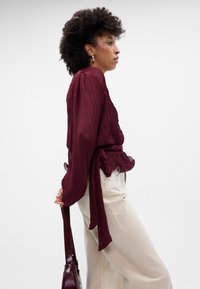 Blusa bordeaux pregueada com mangas longas e uma cintura atada, combinada com calças largas beiges. A modelo segura uma mala escura.