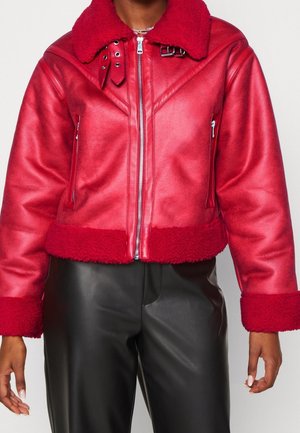 Veste en cuir rouge avec col, poignets et ourlet en peau lainée, fermeture éclair frontale, poches latérales zippées, portée avec un pantalon en cuir noir.