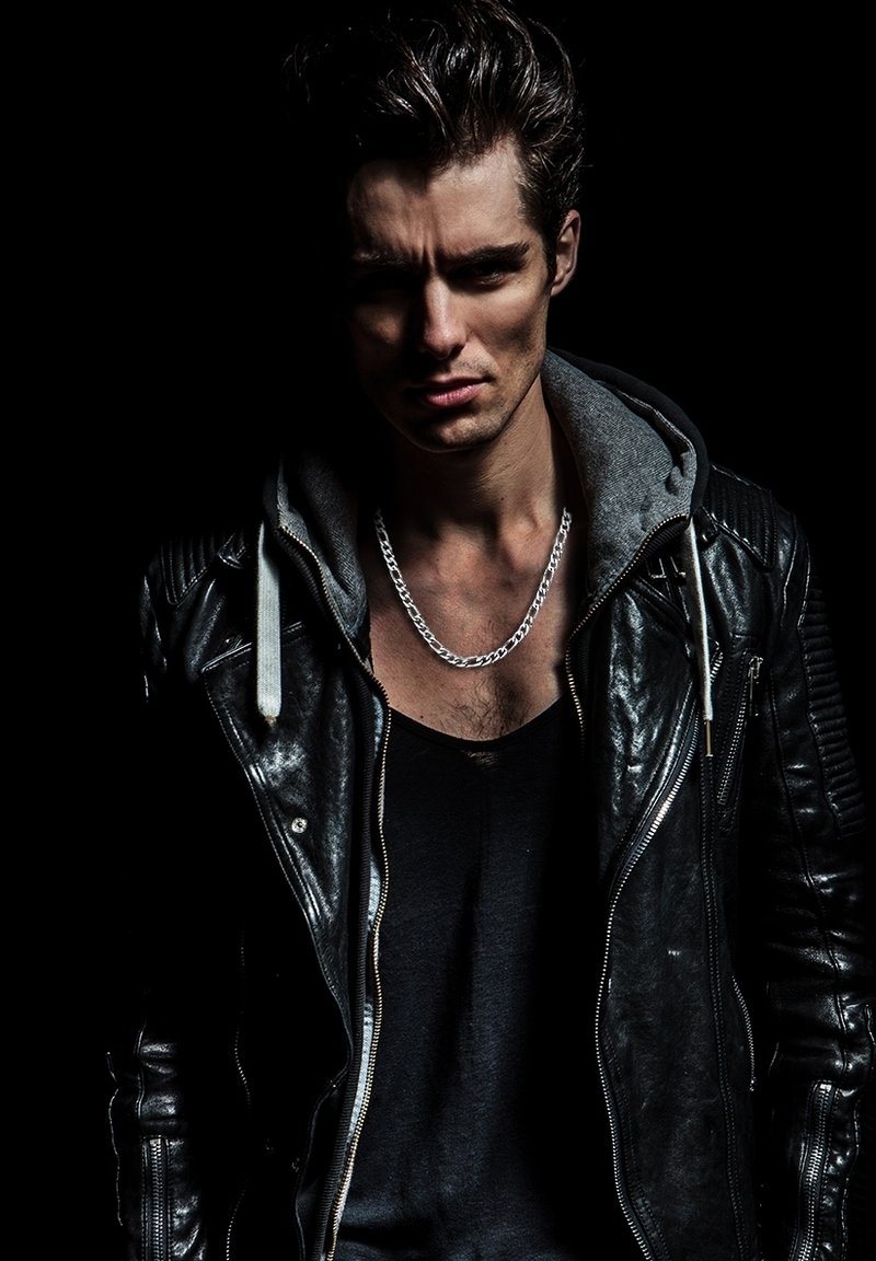 Black leather motorcycle jacket with ribbed shoulders, gray hoodie underneath, silver chain necklace, against a dark background.