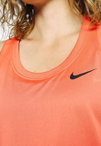 Koralliväriinen urheilullinen toppi, joka on valmistettu sileästä kankaasta. Etupuolella on musta Nike-logo vasemmalla rinnassa ja pyöreä kaula-aukko.