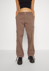 Bruine cargobroek met zijzakken, een elastische tailleband en verstelbare manchetten, gemaakt van textuurstof. Gepaard met zwarte schoenen.