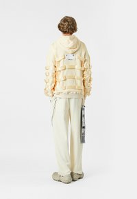 Sudadera de un amarillo pálido con detalle de cinta texturizada y una etiqueta plateada en la parte posterior, combinada con pantalones color crema y zapatillas grises sin cordones.