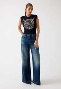 Top sin mangas negro con un gráfico de flores y alas, combinado con jeans de pierna ancha de color azul claro que tienen un acabado desgastado y detalles desgastados.