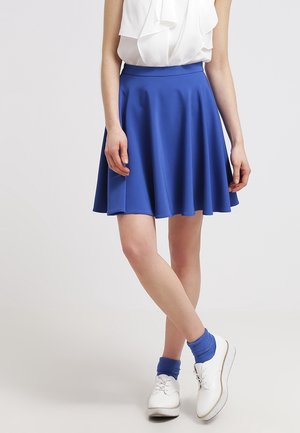 Nainen, joka pukeutunut polvipituiseen kuninkaansiniseen hameeseen, valkoiseen hihattomaan röyhelöiseen puseroon, sinisiin sukkiin ja valkoisiin nyörikenkiin, seisoo jalat ristissä.