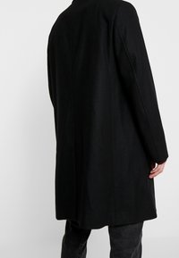 Musta villakangastakki suoran leikkauksen designilla, pitkät hihat ja takakappaleen venttiili. Sileä pinta, minimalistinen tyyli, puettuna tummien farkkujen päällä.