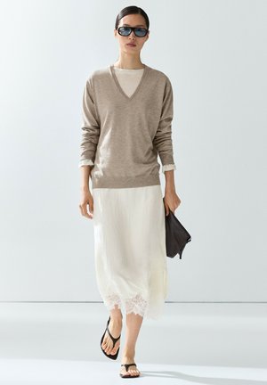 Donna che indossa un maglione beige con scollo a V sopra una gonna bianca con bordo in pizzo, sandali neri, occhiali da sole e tiene una pochette nera mentre cammina in avanti.