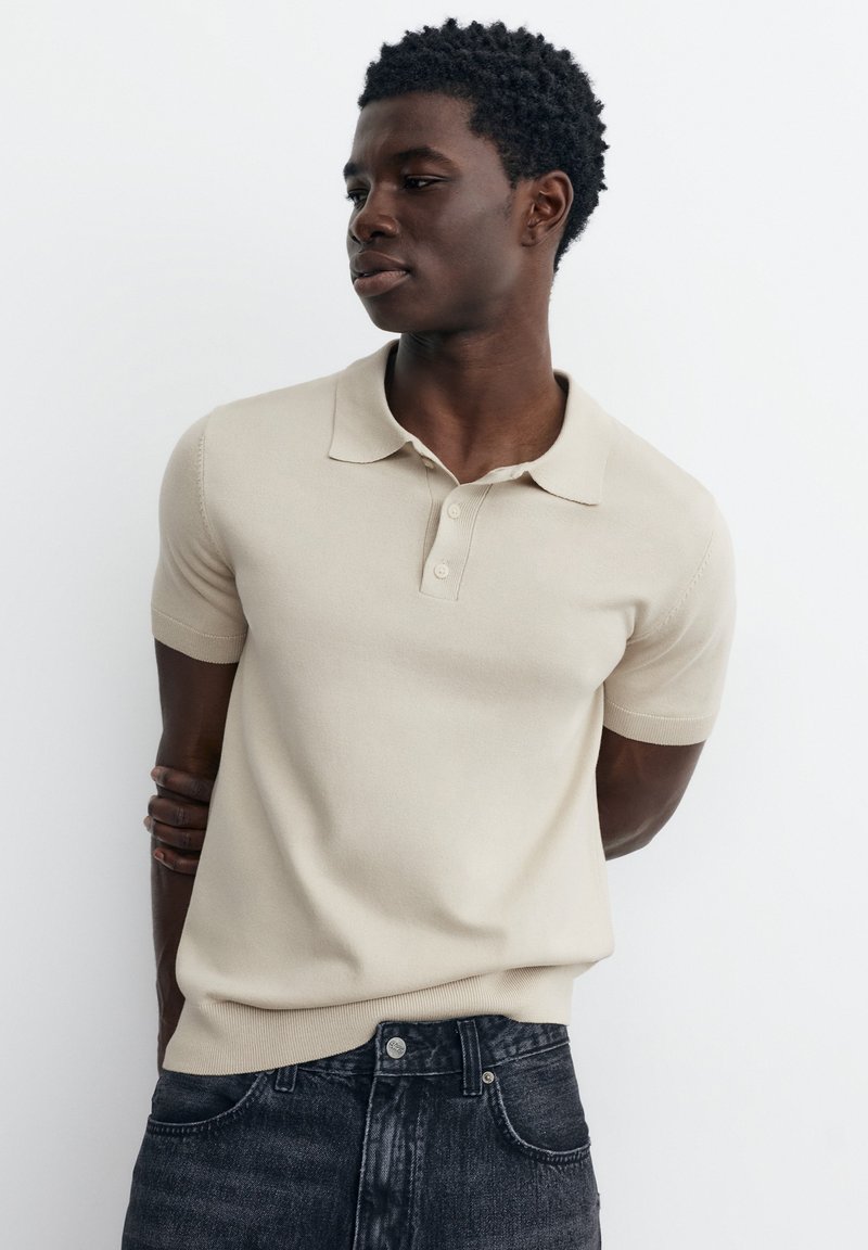 Jeune homme aux cheveux courts et bouclés, portant un polo beige à manches courtes et un jean foncé, debout devant un fond blanc.