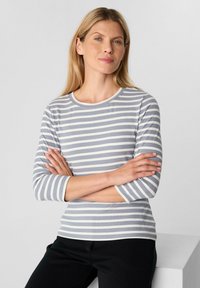 Gestreiftes Langarmhemd in Grau und Weiß, mit rundem Halsausschnitt und gerippter Textur. Das Model posiert mit verschränkten Armen und trägt schwarze Hose.
