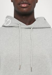 Zbliżenie na osobę w jasnoszarej bluzie z kapturem, z troczkami oraz częściowo widocznymi białymi haftowanymi literami na kapturze.