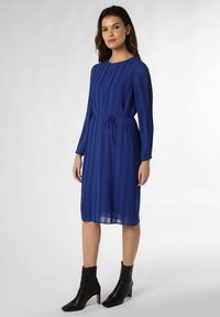 Blaues plissiertes Kleid mit langen Ärmeln, Rundhalsausschnitt und Kordelzug in der Taille. Kombiniert mit schwarzen Stiefeletten, die eine quadratische Zehenform und einen Blockabsatz haben.