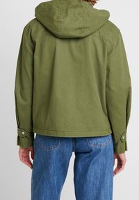 Osoba ubrana w oliwkową, kapturową kurtkę z guzikami przy mankietach oraz niebieskie dżinsy, widziana z tyłu na jednolitym tle.