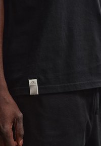 Κοντινό πλάνο ενός ατόμου που φοράει μαύρο πουκάμισο και μαύρο παντελόνι, δείχνοντας μια λευκή ετικέτα με το όνομα της επωνυμίας "Pegador" κοντά στο τελείωμα του πουκαμίσου.
