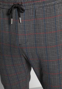 Pantaloni a quadri grigi con cordino nero. Presentano linee rosse e blu nel motivo, evidenziando un design di tessuto testurizzato.