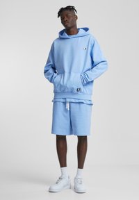 Lichtblauwe hoodie met een kangaroozak, gecombineerd met bijpassende blauwe shorts. Bevat een logopatch en witte veterbanden, gedragen met witte sneakers.