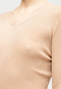 Pull en maille côtelée beige avec un col en V et un tissu texturé, mettant en valeur des lignes verticales et une coupe ajustée.