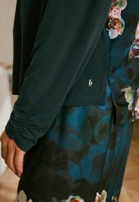 Dunkelgrünes Oberteil mit gerafften Ärmeln, kombiniert mit einem blumengemusterten blauen Rock. Enthält ein kleines silbernes Logo nahe dem Saum.