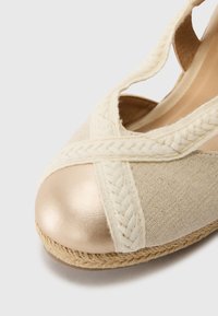 Beige canvas sko med flätad remdesign, metallisk guld tåkapp, och jute-omslagen sula, med en strukturerad yta.