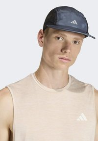 Tmavomodrá čiapka s bočnými sieťovinovými panelmi, rovným šiltom a reflexnými prvkami. Nosí sa s svetlobéžovým tielkom bez rukávov s logom.