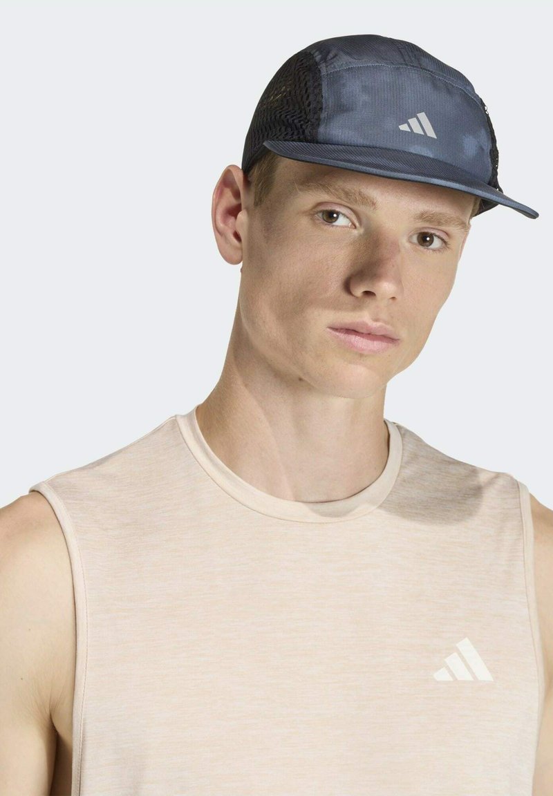 Tmavomodrá čiapka s bočnými sieťovinovými panelmi, rovným šiltom a reflexnými prvkami. Nosí sa s svetlobéžovým tielkom bez rukávov s logom.