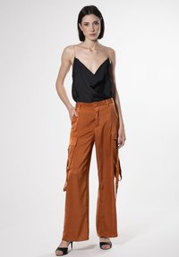 Schwarze Satinhose mit V-Ausschnitt und goldenen Trägern, kombiniert mit orangen weit geschnittenen Hosen mit Cargo-Taschen und seitlichen Bändern, getragen mit schwarzen High Heels.