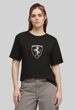 Jeune femme aux longs cheveux auburn portant des boucles d'oreilles anneaux, un t-shirt noir avec un logo de cheval cabré blanc, et un jean gris, posant devant un fond gris.