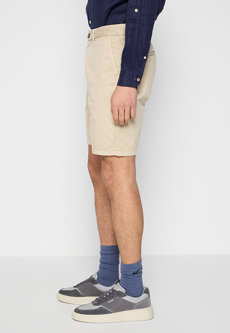 Beżowe szorty wykonane z bawełny, o prostym kroju z bocznymi kieszeniami. W połączeniu z szarymi butami sportowymi i niebieskimi skarpetami w prążki.
