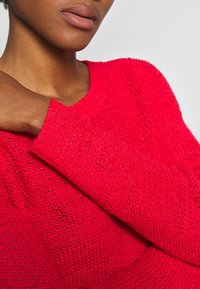 Pull rouge tricoté avec un tissage lâche et un col rond. Comprend des poignets côtelés et un motif texturé. Conçu pour une coupe décontractée.
