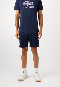 Ναυτικός T-shirt με λογότυπο Lacoste σε ροζ, συνδυασμένο με ναυτικά σορτς. Φορεμένο με λευκά αθλητικά παπούτσια και κάλτσες μέχρι τον αστράγαλο. Χαλαρή αθλητική εμφάνιση.