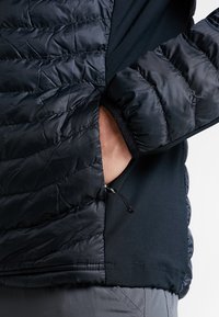 Czarna kurtka puchowa o pikowanej fakturze, z boczną kieszenią i regulowanym dołem. Warstwa pod spodem to jednolity czarny, kontrastowy materiał.