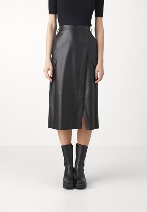 Femme portant une jupe midi en cuir noire avec une fente devant, des poches latérales à rabat, un top noir à manches courtes et des bottines noires sur fond blanc.